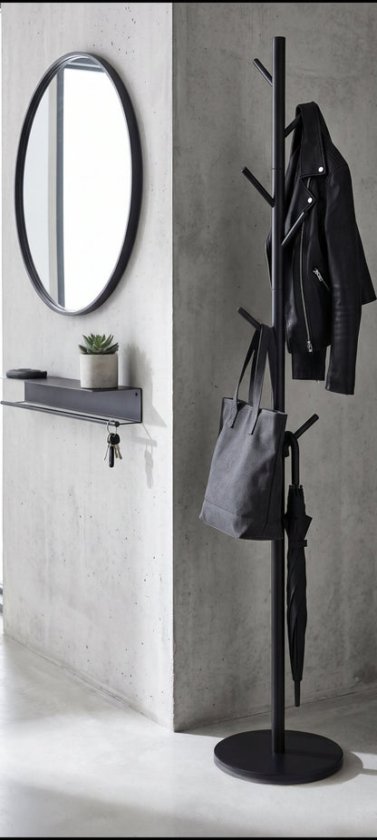 A tall black metal coat rack with multiple hooks stands in a minimalist entryway with a concrete wall, a round black-framed mirror, and a small shelf. A black jacket, gray tote bag, and umbrella are hanging on the rack.