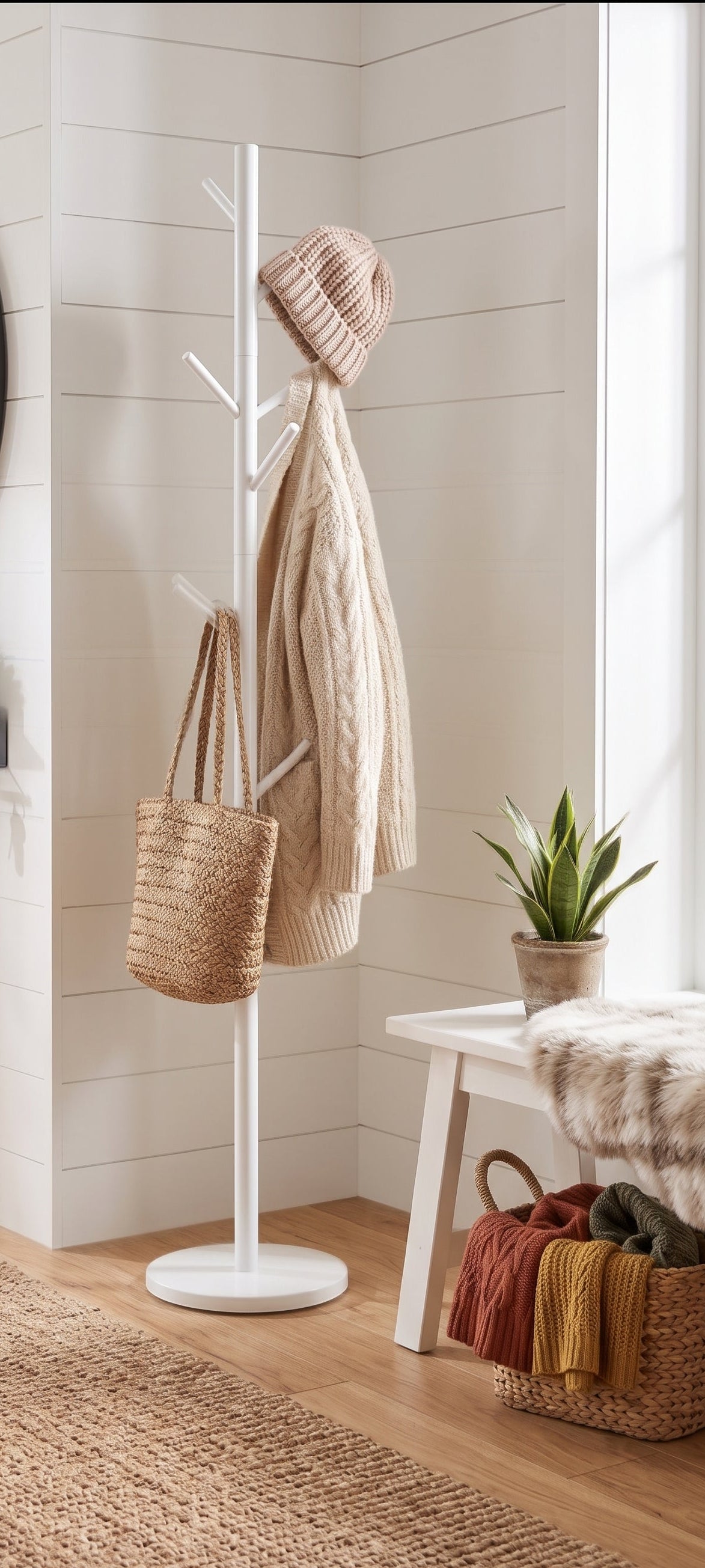 A white floor-mounted coat rack in a bright entryway, holding a beige knit beanie, cream cable-knit cardigan, and a woven straw tote bag. The room has white shiplap walls, a wood floor, a small white bench, and a woven rug.